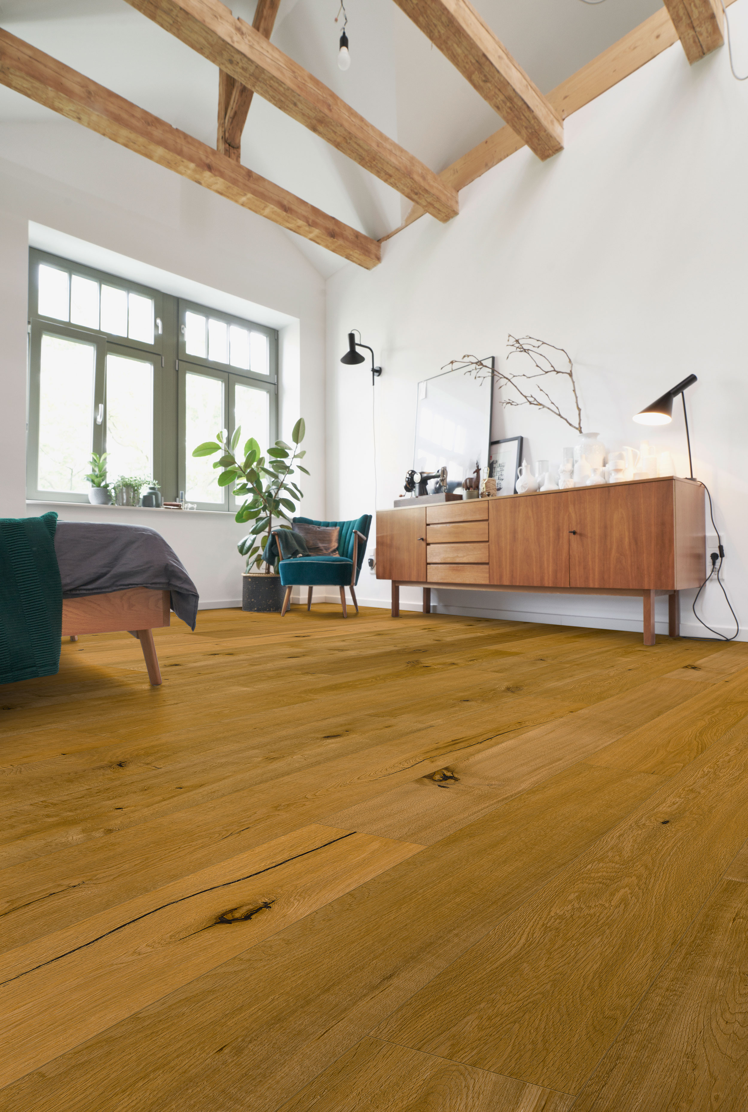 Modernes Schlafzimmer mit Holzfußboden, sichtbaren Holzbalken an der Decke, großem Fenster, Bett mit dunkler Bettwäsche, grünem Sessel und Holzkommode mit Dekoration.